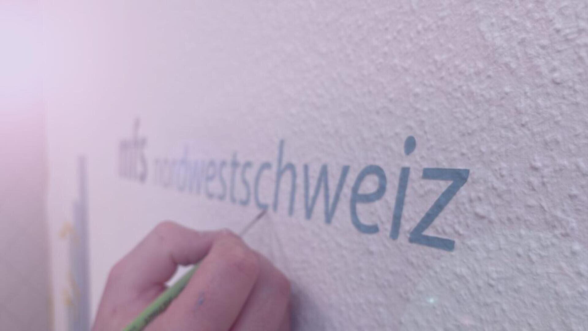 Eine Hand malt das Wort „Nordwestschweiz“ in blauen Buchstaben auf eine strukturierte weiße Wand; aus der linken oberen Ecke strahlt ein sanftes rosa Licht.