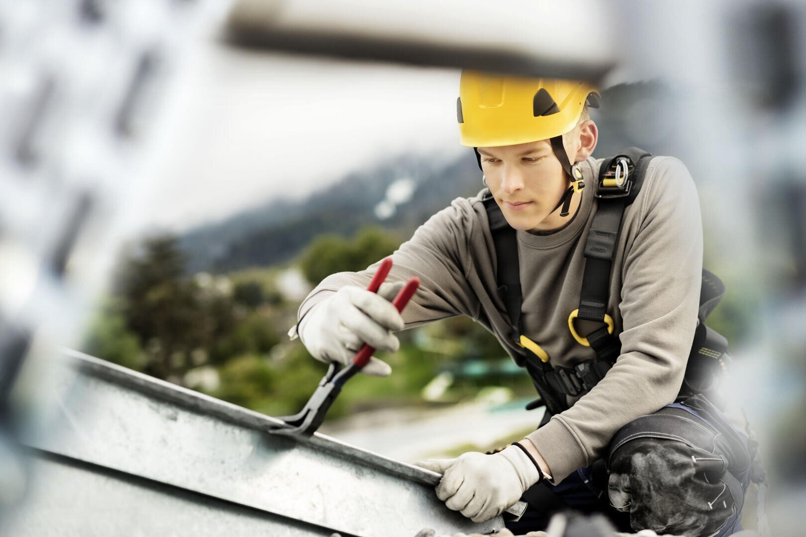 Ein Bauarbeiter mit gelbem Schutzhelm und Sicherheitsgurt crimpt mit einer Zange eine Metalldachplatte.