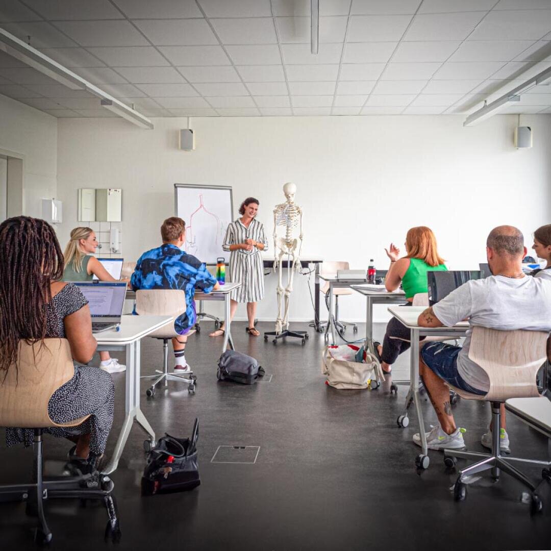 Ein Lehrer steht neben einem Skelettmodell und einer Whiteboard-Tafel und hält den Schülern, die in einem hellen Klassenzimmer an ihren Schreibtischen sitzen und Laptops vor sich haben, einen Vortrag.
