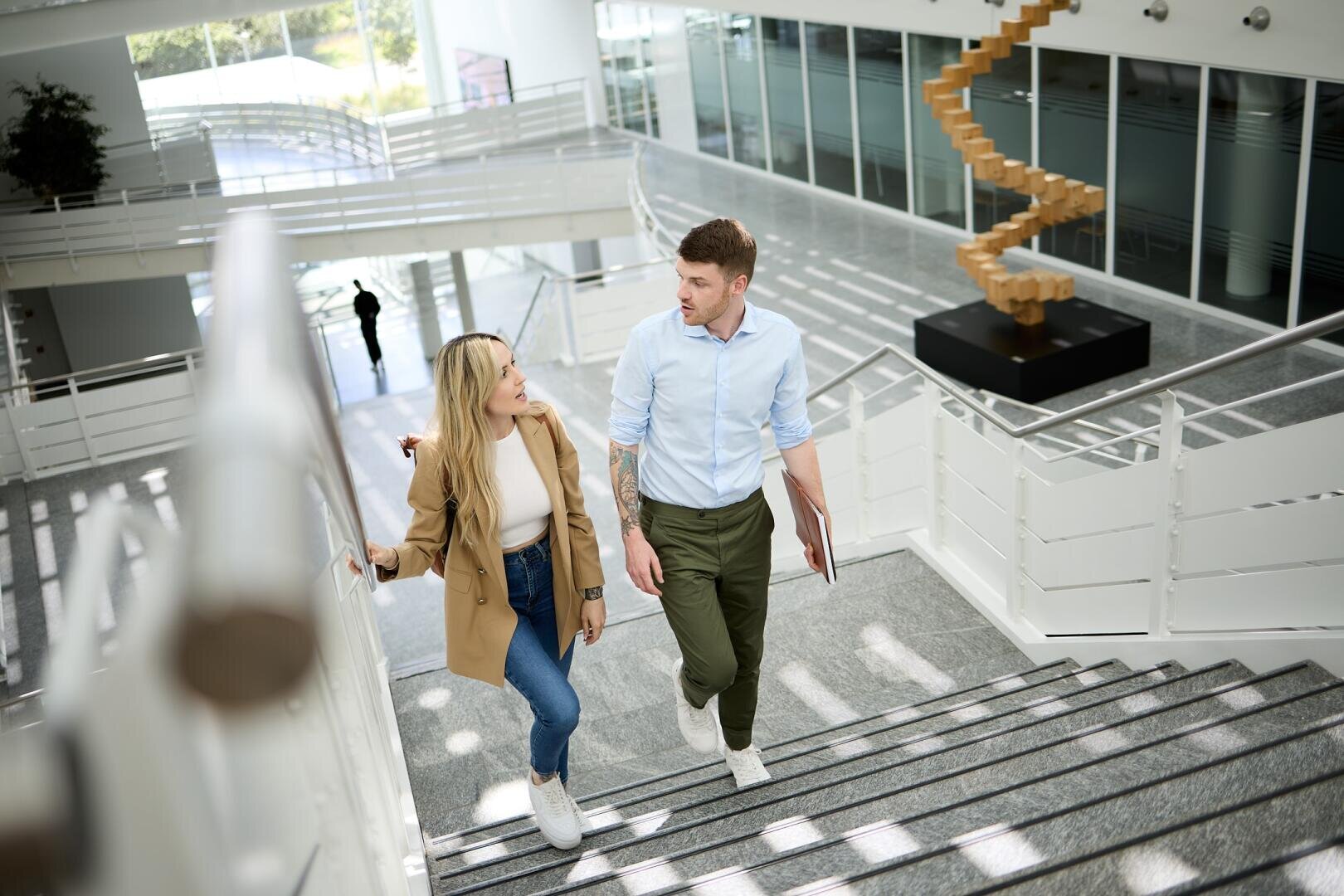 Zwei Personen, eine Frau und ein Mann, gehen eine Treppe im Inneren eines modernen Gebäudes mit großen Fenstern und einer Spiralskulptur im Hintergrund hinauf. Sie scheinen ein Gespräch zu führen.