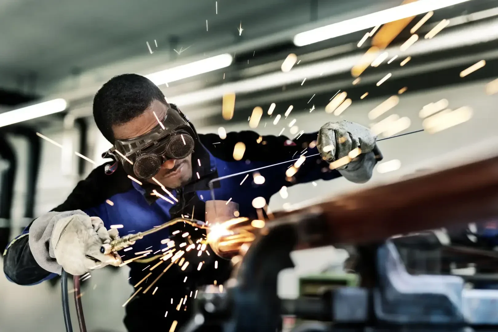 Eine Person, die eine Schutzbrille und Handschuhe trägt, schweißt mit einem Werkzeug Metall und verursacht dabei helle, umherfliegende Funken in einer Industriewerkstatt.