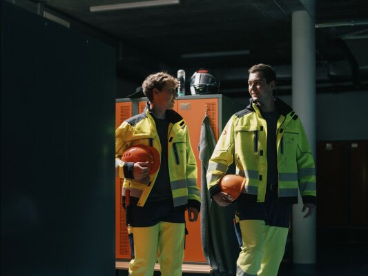 Zwei Personen in gelben Sicherheitswesten und dunklen Hosen halten orangefarbene Helme in den Händen und gehen lächelnd durch einen Umkleideraum in einem Industriebetrieb, im Hintergrund sind Spinde und Schutzausrüstung zu sehen.