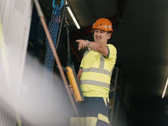 Ein Bauarbeiter, der einen Helm und eine Warnweste trägt, steht auf einer Baustelle, hält ein Baugerät in der Hand, lächelt und zeigt nach vorne.