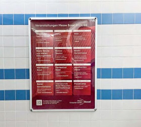 Auf einer hellblau-weiss gekachelten Wand ist ein rot-weisses Veranstaltungsplakat in deutscher Sprache angebracht, das verschiedene Veranstaltungen der Messe Basel für 2022/23 auflistet. Das Plakat ist in einem Metallrahmen befestigt.
