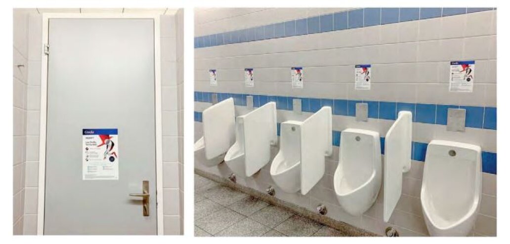 Eine Toilette mit einer geschlossenen weißen Tür auf der linken Seite und fünf weißen Pissoirs an einer gefliesten Wand auf der rechten Seite, über denen jeweils ein Schild angebracht ist. Blaue Akzentfliesen verlaufen horizontal an der Wand entlang.